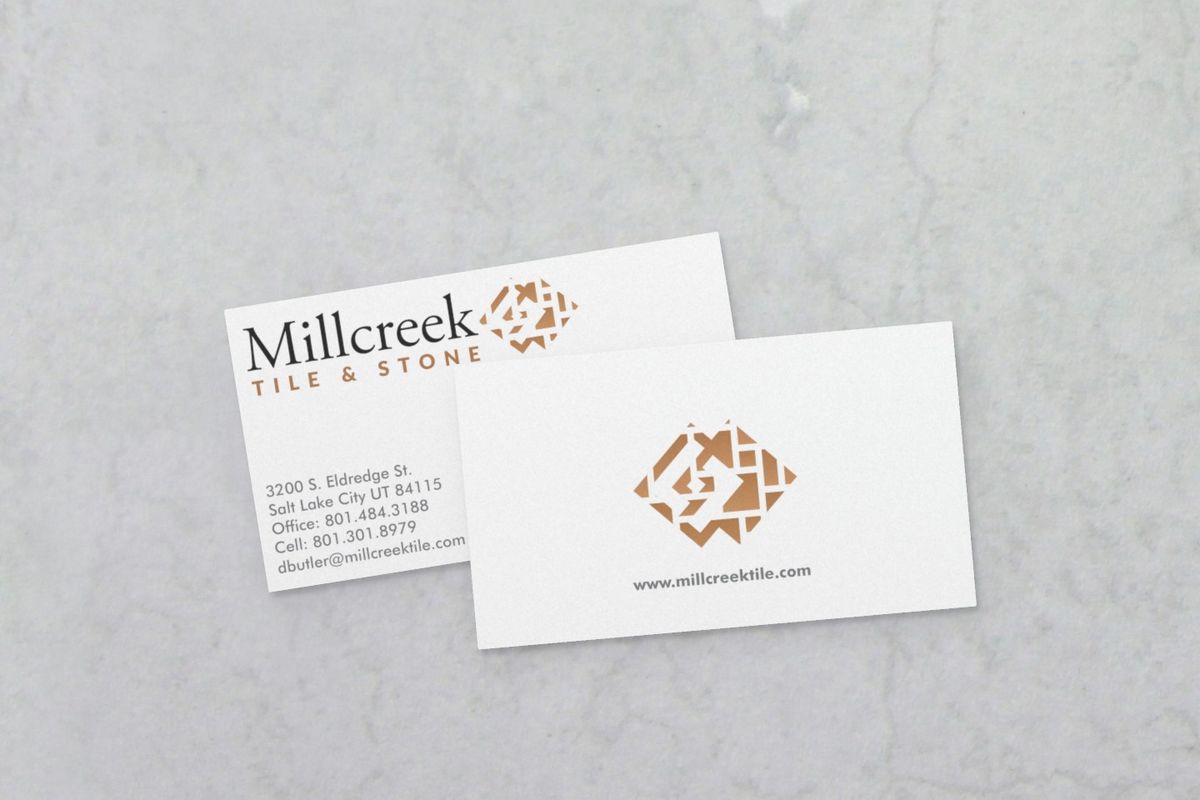 Two white Millcreek Tile & Stone business cards on gray marble: one with the logo, street address, phone numbers and email, partly covered by the other, which shows the copper-colored tile logo and web address.