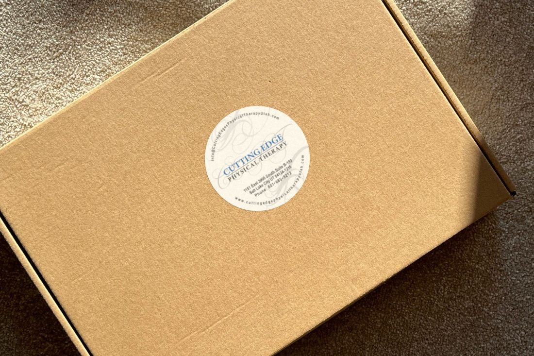 Round white Cutting Edge Physical Therapy label with blue and gray lettering and contact details, applied to the lid of a brown shipping box.