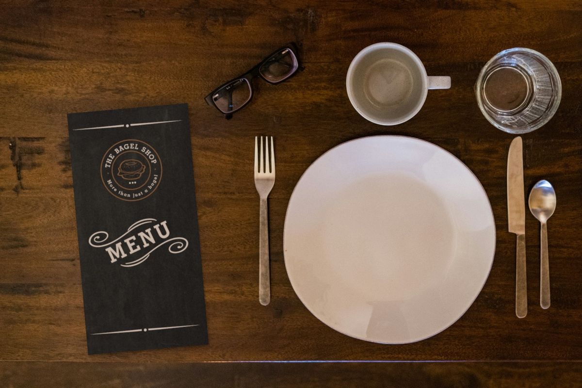 The Bagel Shop menu, a tall card with a chalkboard-style cover showing the round logo and "Menu" in white lettering, beside a place setting on a dark wooden table.