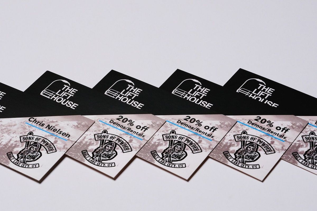 Overlapping rows of printed promotional cards: black cards with a white chairlift logo, and light cards over a snowy mountain photo offering 20% off demos and rentals.