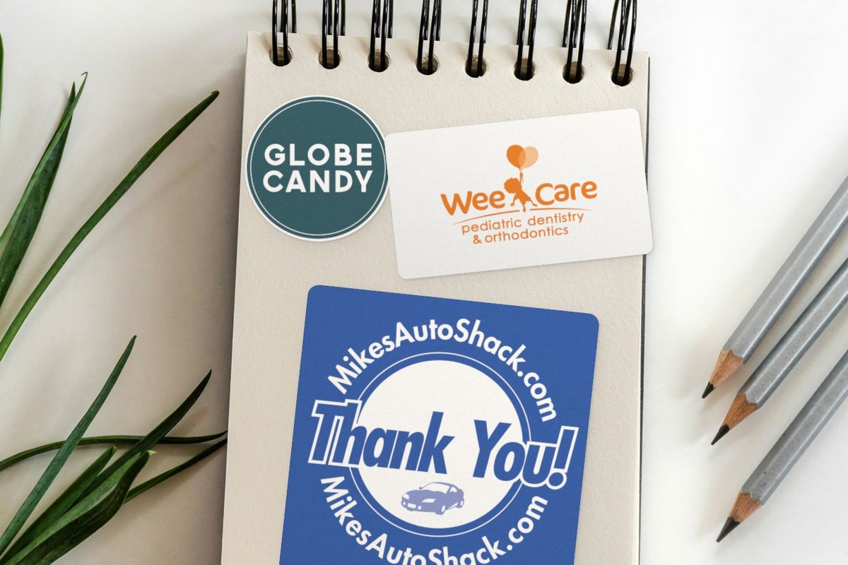 Five logo stickers on a white desk: rounded rectangles for Gonzalez Group and Wright Dental Group, and on a spiral notepad a round Globe Candy sticker, a Wee Care pediatric dentistry rectangle and a square blue "Thank You!" sticker for MikesAutoShack.com.