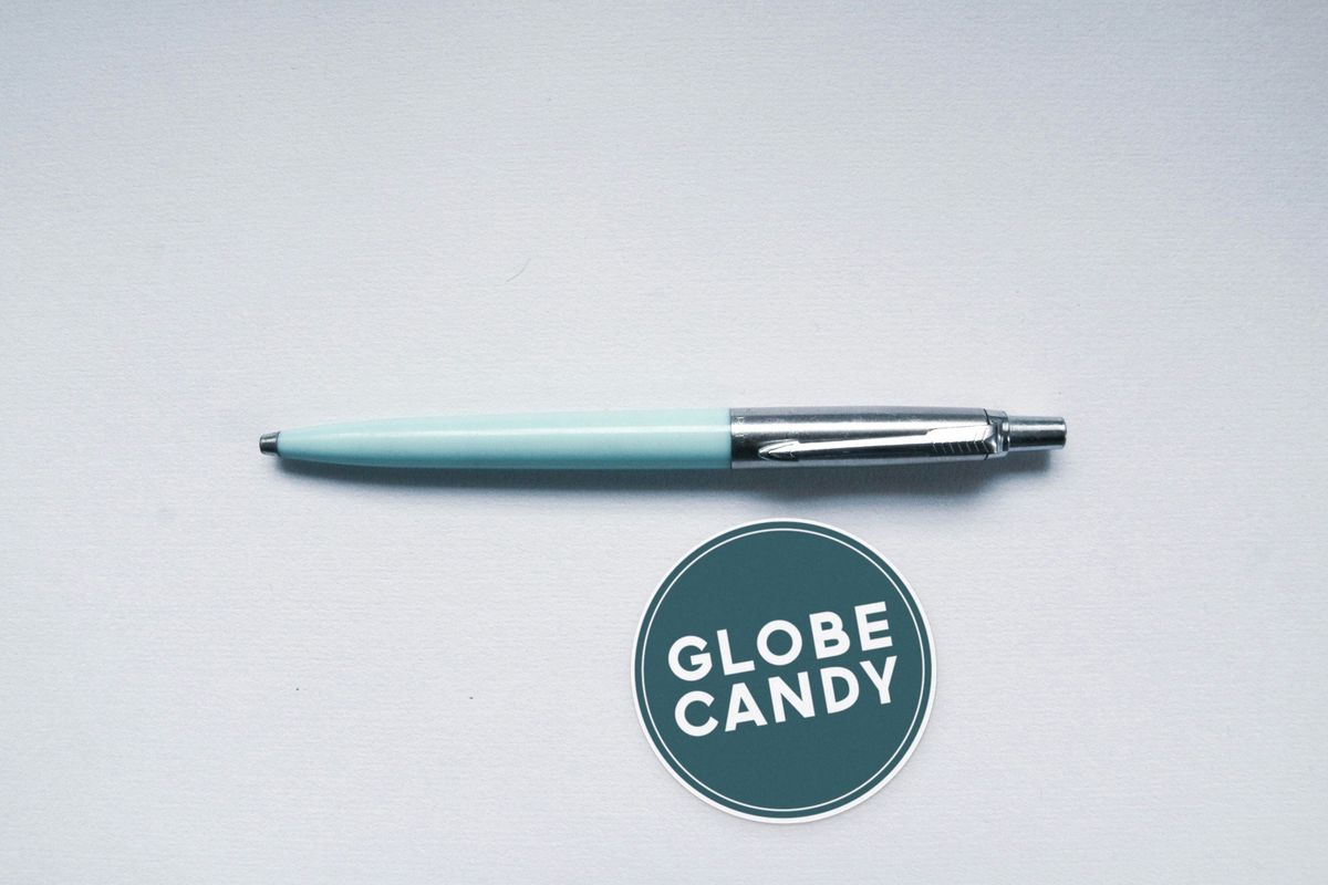 Round dark teal Globe Candy logo sticker with white lettering, on a pale gray surface below a mint-green pen.